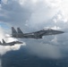 Two U.S. Air Force F-15E Strike Eagle fighter jets are flying in formation above cloud cover. The lead aircraft displays 'SJ' tail codes, identifying it as belonging to the 4th Fighter Wing based at Seymour Johnson Air Force Base, North Carolina. Both aircraft appear to be carrying external ordnance and fuel configurations on their hardpoints.