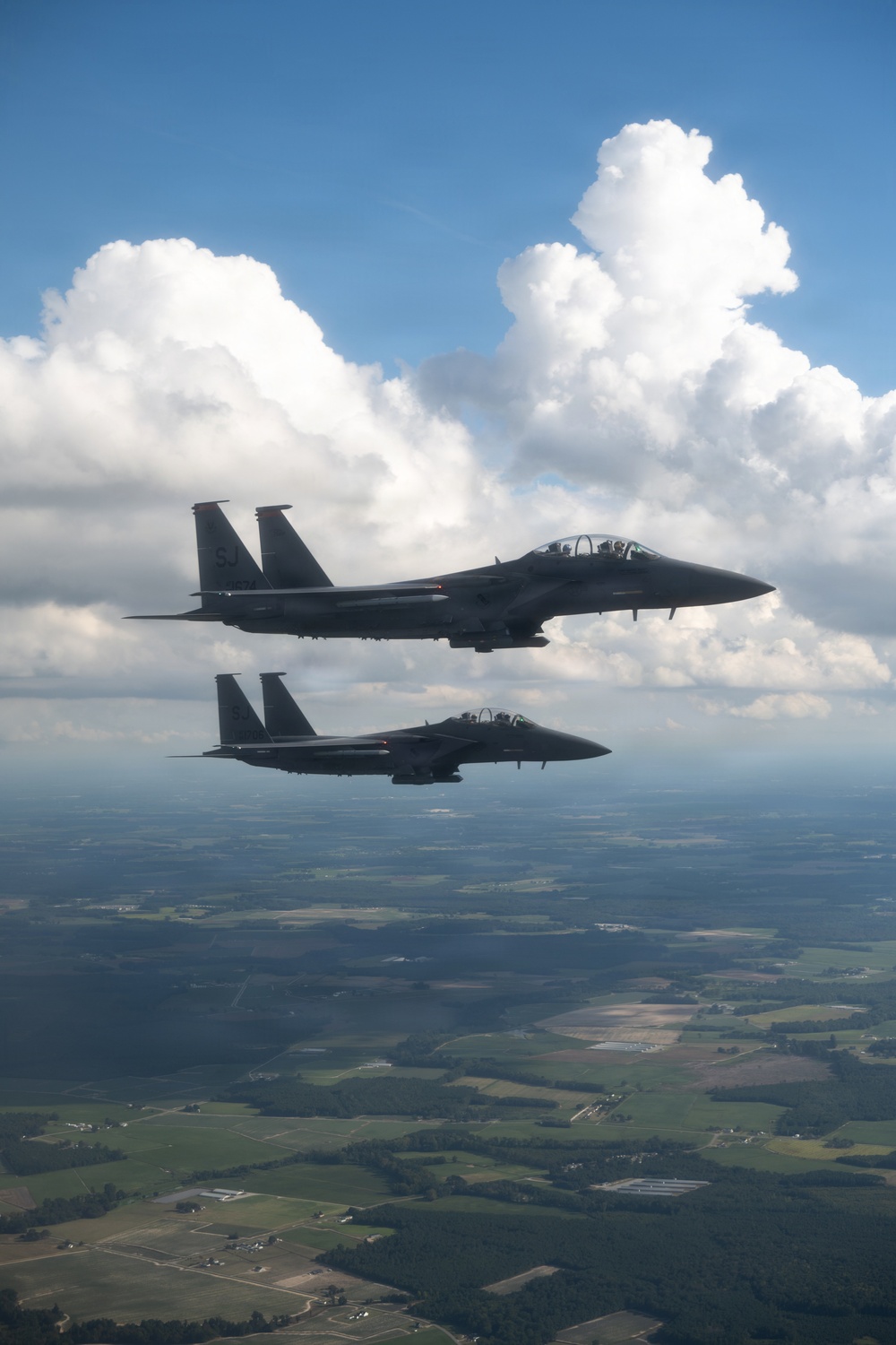 Two dark-colored military fighter jets, identifiable as F-15 Eagles, are flying in formation over a rural landscape. The aircraft are photographed from a slightly elevated angle, with large cumulus clouds visible in the background. The tail code 'SJ' is visible on the lead aircraft, indicating assignment to Seymour Johnson Air Force Base.