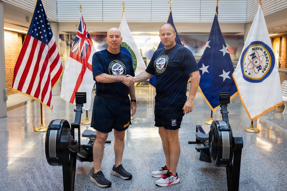 Two men in matching navy blue 'Naval Reactors' t-shirts and athletic shorts shake hands between two rowing machines (Concept2 ergometers) in what appears to be an official building lobby. Behind them, multiple flags are displayed including the United States flag, the British White Ensign (Royal Navy), a blue flag with stars, and a white 'Naval Reactors' (NR) organizational flag. The setting and attire suggest a formal athletic or fitness challenge event related to the Naval Reactors program.