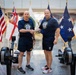 Two men in matching navy blue 'Naval Reactors' t-shirts and athletic shorts shake hands between two rowing machines (Concept2 ergometers) in what appears to be an official building lobby. Behind them, multiple flags are displayed including the United States flag, the British White Ensign (Royal Navy), a blue flag with stars, and a white 'Naval Reactors' (NR) organizational flag. The setting and attire suggest a formal athletic or fitness challenge event related to the Naval Reactors program.