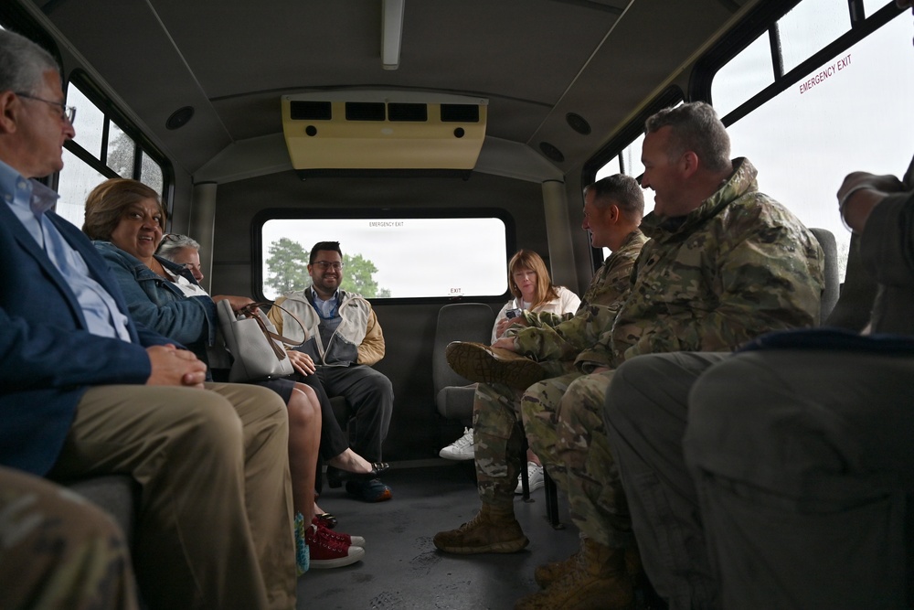 A group of civilians and two military personnel in camouflage uniforms are seated inside a small bus or shuttle vehicle, engaged in conversation. The group appears to be on a tour or transport, with people seated along both sides of the aisle facing each other. The atmosphere appears relaxed and social, with several individuals smiling.