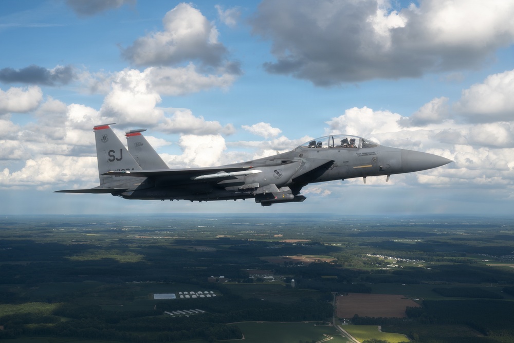A U.S. Air Force F-15E Strike Eagle fighter jet is photographed in flight over a rural, forested landscape under a partly cloudy sky. The aircraft bears the tail code 'SJ', indicating it is assigned to Seymour Johnson Air Force Base in North Carolina. The two-seat aircraft appears to be on a training or transit mission at medium altitude.