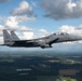 A U.S. Air Force F-15E Strike Eagle fighter jet is photographed in flight over a rural, forested landscape under a partly cloudy sky. The aircraft bears the tail code 'SJ', indicating it is assigned to Seymour Johnson Air Force Base in North Carolina. The two-seat aircraft appears to be on a training or transit mission at medium altitude.
