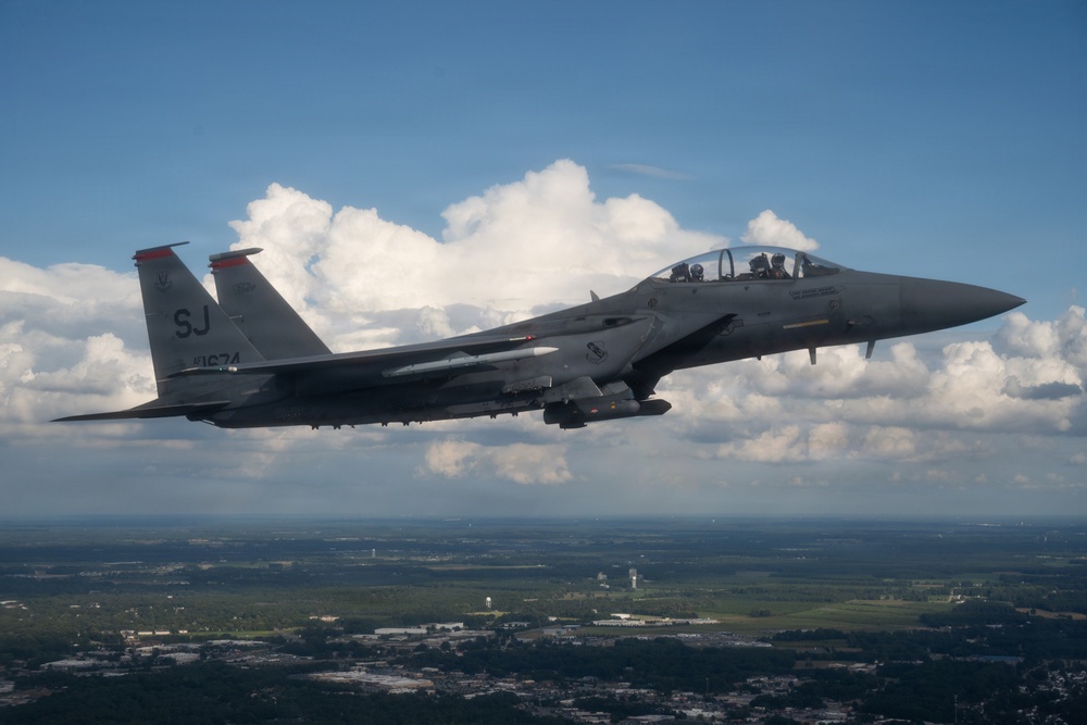A U.S. Air Force F-15E Strike Eagle fighter jet is photographed in flight over a green, partially developed landscape. The twin-seat aircraft bears the tail code 'SJ' indicating assignment to Seymour Johnson Air Force Base in North Carolina. The aircraft appears to be carrying external stores and has two crew members visible in the cockpit.