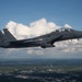 A U.S. Air Force F-15E Strike Eagle fighter jet is photographed in flight over a green, partially developed landscape. The twin-seat aircraft bears the tail code 'SJ' indicating assignment to Seymour Johnson Air Force Base in North Carolina. The aircraft appears to be carrying external stores and has two crew members visible in the cockpit.