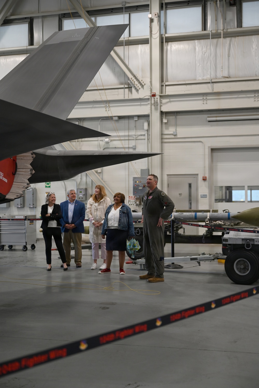 A military briefing or tour is taking place inside an aircraft hangar, where a person in a flight suit appears to be presenting to a small group of civilians. The group is standing beneath the tail section of what appears to be an F-35 stealth fighter jet. A barrier tape in the foreground reads '134th Fighter Wing,' indicating this is a National Guard or Air Force installation.