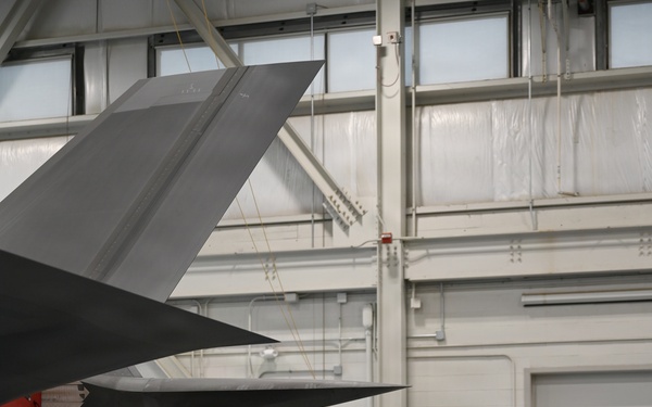 A military briefing or tour is taking place inside an aircraft hangar, where a person in a flight suit appears to be presenting to a small group of civilians. The group is standing beneath the tail section of what appears to be an F-35 stealth fighter jet. A barrier tape in the foreground reads '134th Fighter Wing,' indicating this is a National Guard or Air Force installation.