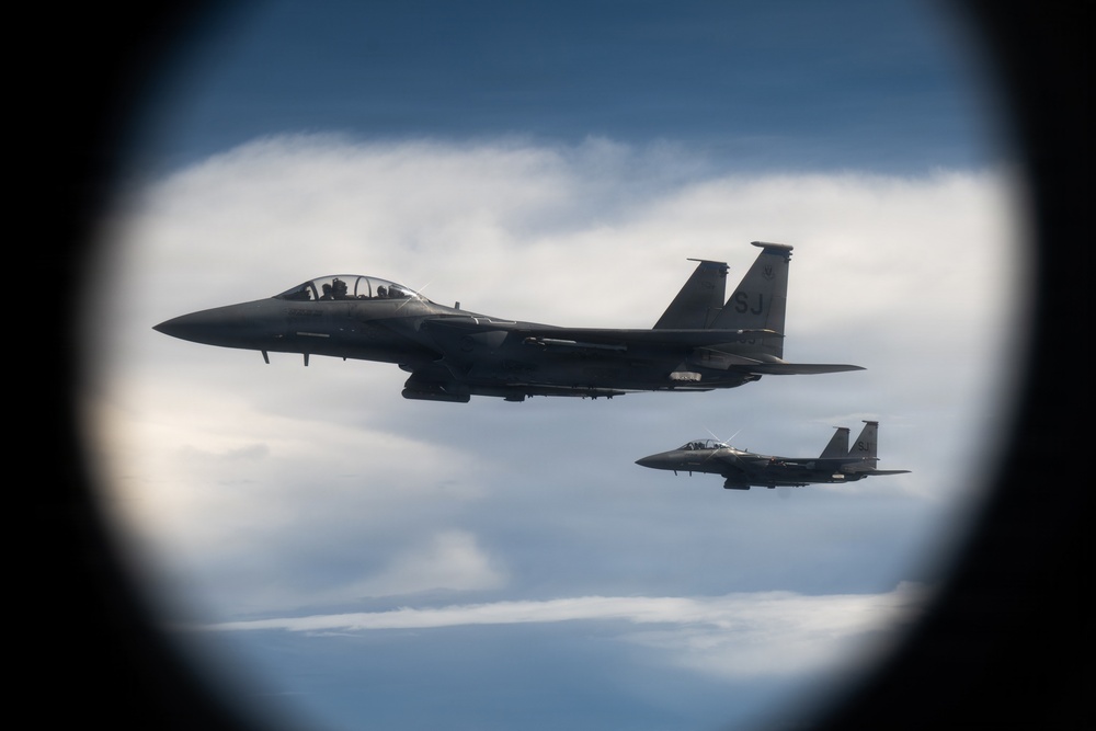 Two military fighter jets, identifiable as F-15 Eagles, are photographed in flight through what appears to be a porthole or oval window of another aircraft. The lead aircraft displays 'SJ' tail codes on its twin vertical stabilizers, indicating assignment to Seymour Johnson Air Force Base. Both aircraft are flying in formation against a partly cloudy sky.