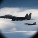 Two military fighter jets, identifiable as F-15 Eagles, are photographed in flight through what appears to be a porthole or oval window of another aircraft. The lead aircraft displays 'SJ' tail codes on its twin vertical stabilizers, indicating assignment to Seymour Johnson Air Force Base. Both aircraft are flying in formation against a partly cloudy sky.