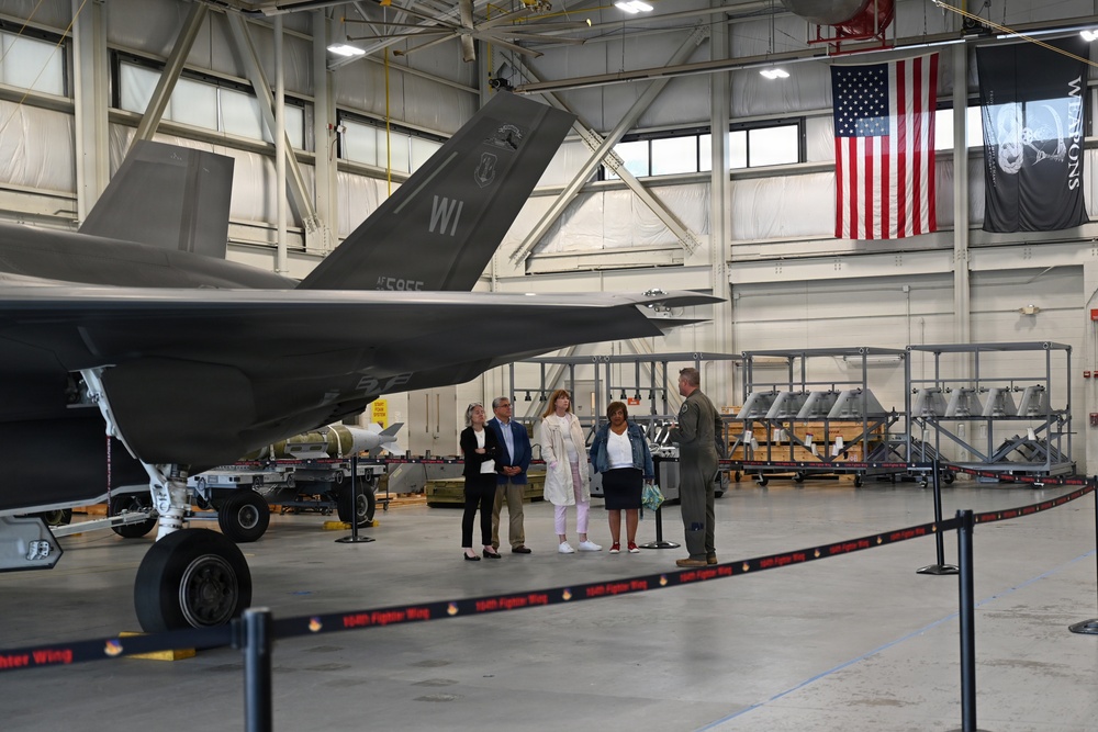 A military briefing or tour is taking place inside an aircraft hangar, where a uniformed pilot or officer is addressing a small group of four civilians. A stealth fighter jet, identifiable as an F-35 with 'WI' tail markings and serial number AF595F, is prominently displayed in the foreground. The hangar also displays an American flag, a 'WEAPONS' banner, and racks of munitions/ordnance in the background.