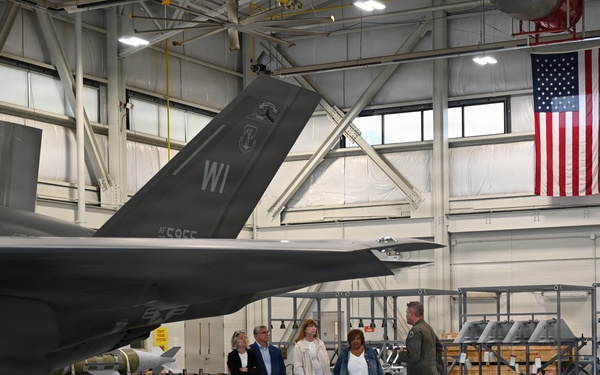 A military briefing or tour is taking place inside an aircraft hangar, where a uniformed pilot or officer is addressing a small group of four civilians. A stealth fighter jet, identifiable as an F-35 with 'WI' tail markings and serial number AF595F, is prominently displayed in the foreground. The hangar also displays an American flag, a 'WEAPONS' banner, and racks of munitions/ordnance in the background.