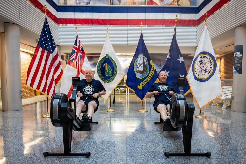 Two men in matching navy blue 'Naval Reactors' t-shirts are using rowing machines (Concept2 ergometers) in what appears to be an official government or military building lobby. They are positioned in front of an array of flags including the United States flag, a United Kingdom flag, the United States Navy flag, a Department of Energy flag, and a Naval Reactors flag. The setting suggests an official fitness event or challenge at a Naval Reactors facility.