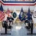 Two men in matching navy blue 'Naval Reactors' t-shirts are using rowing machines (Concept2 ergometers) in what appears to be an official government or military building lobby. They are positioned in front of an array of flags including the United States flag, a United Kingdom flag, the United States Navy flag, a Department of Energy flag, and a Naval Reactors flag. The setting suggests an official fitness event or challenge at a Naval Reactors facility.