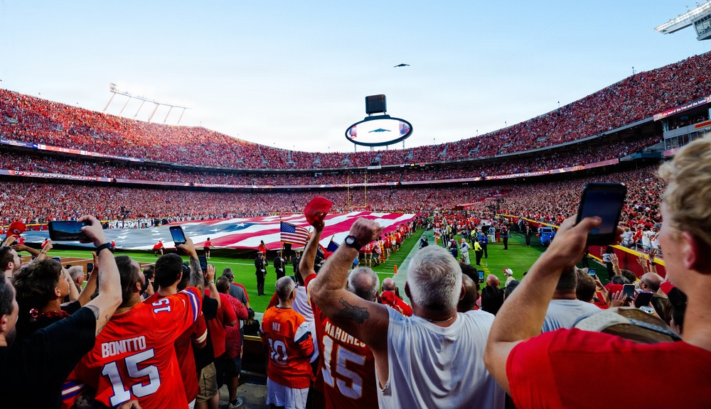 A packed NFL stadium during a pre-game ceremony featuring a large American flag spread across the field, likely during the national anthem. Fans dressed predominantly in red and orange jerseys are standing, cheering, and recording the moment on their phones. A military flyover aircraft is visible in the sky above the stadium.