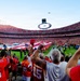 A packed NFL stadium during a pre-game ceremony featuring a large American flag spread across the field, likely during the national anthem. Fans dressed predominantly in red and orange jerseys are standing, cheering, and recording the moment on their phones. A military flyover aircraft is visible in the sky above the stadium.