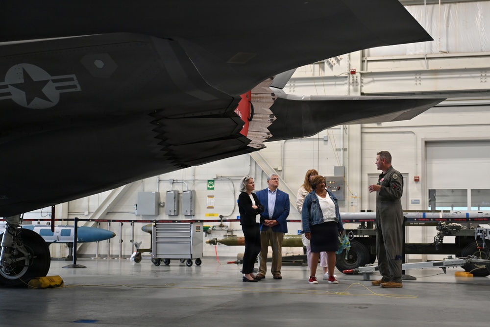 A military personnel member in flight suit is briefing a group of four civilians inside an aircraft hangar. They are standing beneath the underside of what appears to be an F-35 stealth fighter jet, which displays a US military star insignia. The hangar setting includes visible maintenance equipment, munitions, and tool carts in the background.