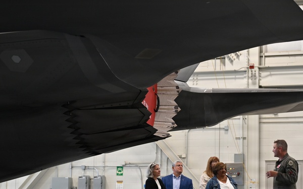 A military personnel member in flight suit is briefing a group of four civilians inside an aircraft hangar. They are standing beneath the underside of what appears to be an F-35 stealth fighter jet, which displays a US military star insignia. The hangar setting includes visible maintenance equipment, munitions, and tool carts in the background.