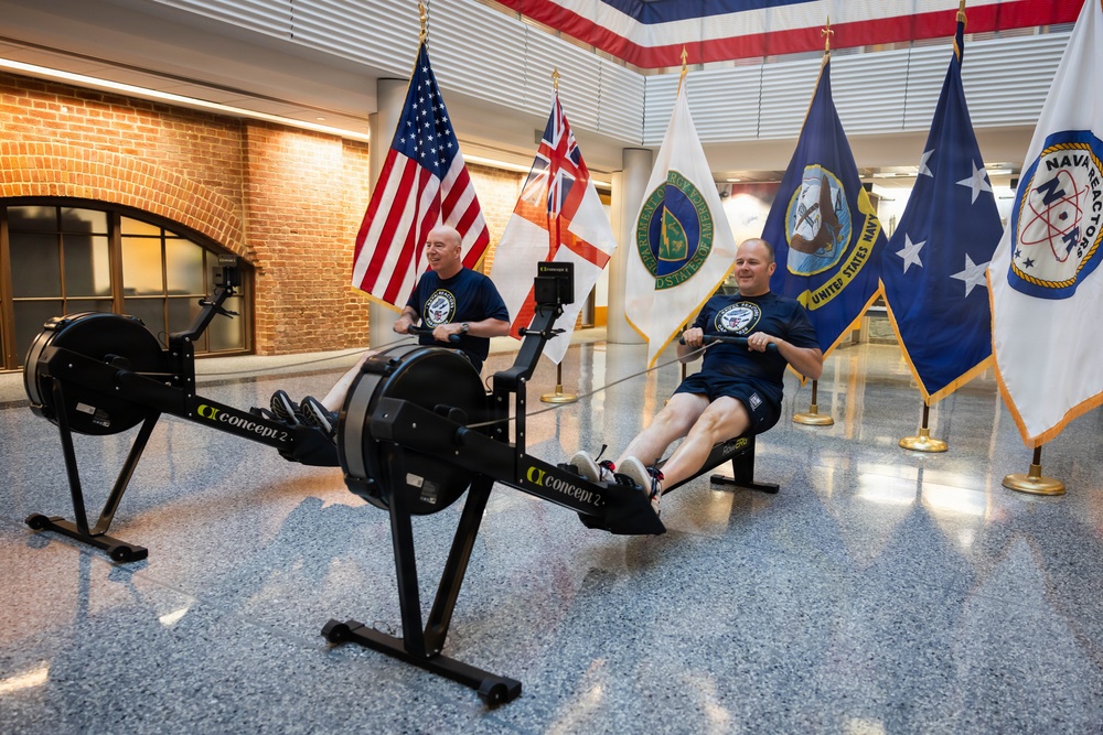 Two men in matching navy blue t-shirts are using Concept2 rowing machines in what appears to be an official military building lobby. Behind them, several flags are displayed including the United States flag, a British Royal Navy White Ensign, a United States Navy flag, and a Naval Reactors flag. The setting suggests a formal military or government facility, likely during a fitness event or challenge.