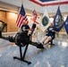 Two men in matching navy blue t-shirts are using Concept2 rowing machines in what appears to be an official military building lobby. Behind them, several flags are displayed including the United States flag, a British Royal Navy White Ensign, a United States Navy flag, and a Naval Reactors flag. The setting suggests a formal military or government facility, likely during a fitness event or challenge.