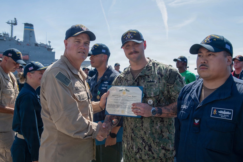 A U.S. Navy award ceremony is taking place outdoors, likely at a naval pier or port. A senior Navy officer in tan uniform is shaking hands with a sailor in digital camouflage (NWU Type III) while both hold a Department of the Navy certificate, identified as a Navy and Marine Corps Commendation Medal citation. A group of Navy personnel in various uniforms are gathered in the background near a large naval vessel.