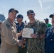 A U.S. Navy award ceremony is taking place outdoors, likely at a naval pier or port. A senior Navy officer in tan uniform is shaking hands with a sailor in digital camouflage (NWU Type III) while both hold a Department of the Navy certificate, identified as a Navy and Marine Corps Commendation Medal citation. A group of Navy personnel in various uniforms are gathered in the background near a large naval vessel.