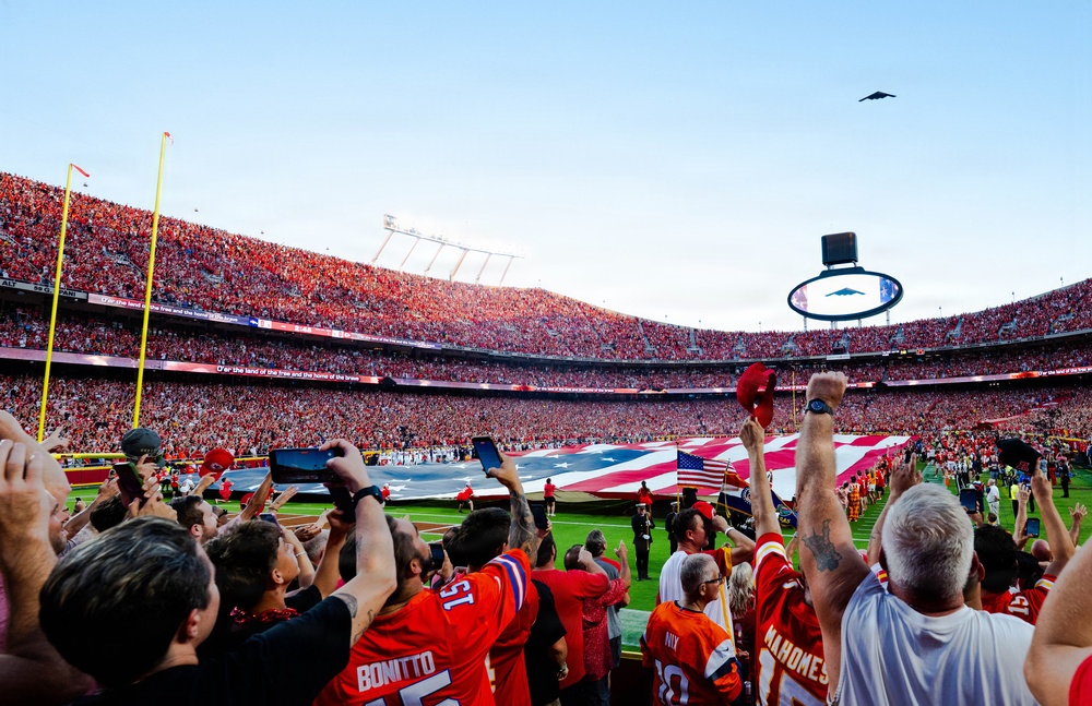 A packed NFL stadium filled predominantly with fans wearing red Kansas City Chiefs gear during a pre-game ceremony. A large American flag is stretched across the field while a military flyover aircraft — appearing to be a B-2 Spirit stealth bomber — passes overhead. Fans in the foreground are cheering, raising their arms, and capturing the moment on phones and tablets.