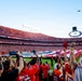 A packed NFL stadium filled predominantly with fans wearing red Kansas City Chiefs gear during a pre-game ceremony. A large American flag is stretched across the field while a military flyover aircraft — appearing to be a B-2 Spirit stealth bomber — passes overhead. Fans in the foreground are cheering, raising their arms, and capturing the moment on phones and tablets.