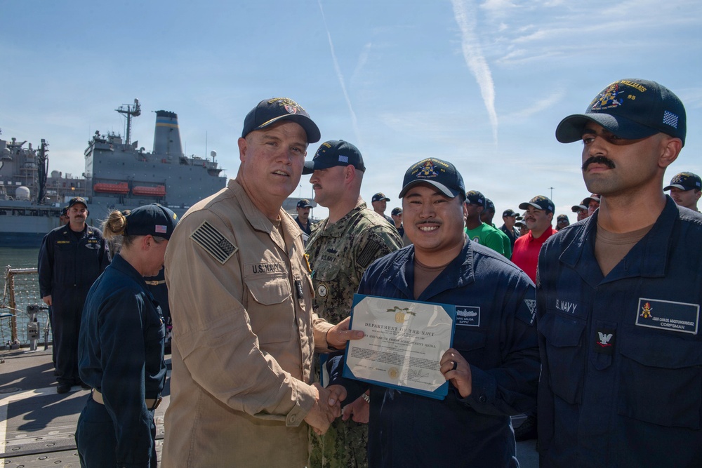 A U.S. Navy officer in tan/khaki uniform is presenting a Department of the Navy certificate to a sailor in navy blue coveralls on what appears to be a ship's deck or pier. The ceremony is taking place outdoors with a group of assembled sailors watching in the background. A large naval vessel is visible in the background.