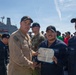A U.S. Navy officer in tan/khaki uniform is presenting a Department of the Navy certificate to a sailor in navy blue coveralls on what appears to be a ship's deck or pier. The ceremony is taking place outdoors with a group of assembled sailors watching in the background. A large naval vessel is visible in the background.