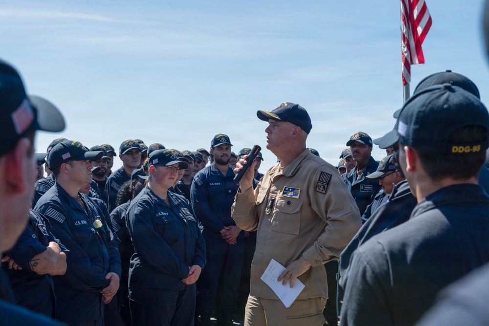 A senior U.S. Navy officer in tan/khaki uniform addresses a large group of enlisted sailors wearing dark blue Navy working uniforms outdoors. The gathering appears to be an all-hands or formation briefing taking place on what is likely a ship's deck or pier. An American flag is visible in the background on a flagpole.