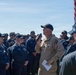 A senior U.S. Navy officer in tan/khaki uniform addresses a large group of enlisted sailors wearing dark blue Navy working uniforms outdoors. The gathering appears to be an all-hands or formation briefing taking place on what is likely a ship's deck or pier. An American flag is visible in the background on a flagpole.