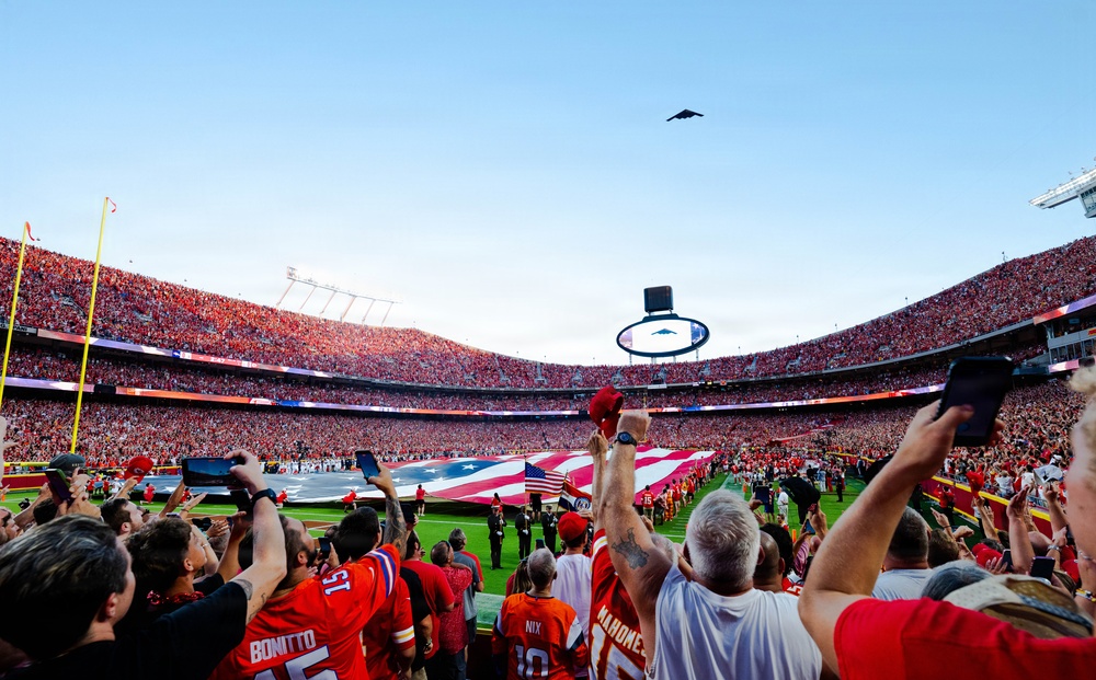 A packed NFL stadium during a pre-game ceremony featuring a large American flag spread across the field and a military flyover by what appears to be a B-2 stealth bomber. Fans in red and orange jerseys fill the stands and foreground, many holding up phones and tablets to capture the moment. The scene is consistent with a Kansas City Chiefs home game based on visible jersey names and team colors.