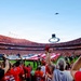 A packed NFL stadium during a pre-game ceremony featuring a large American flag spread across the field and a military flyover by what appears to be a B-2 stealth bomber. Fans in red and orange jerseys fill the stands and foreground, many holding up phones and tablets to capture the moment. The scene is consistent with a Kansas City Chiefs home game based on visible jersey names and team colors.