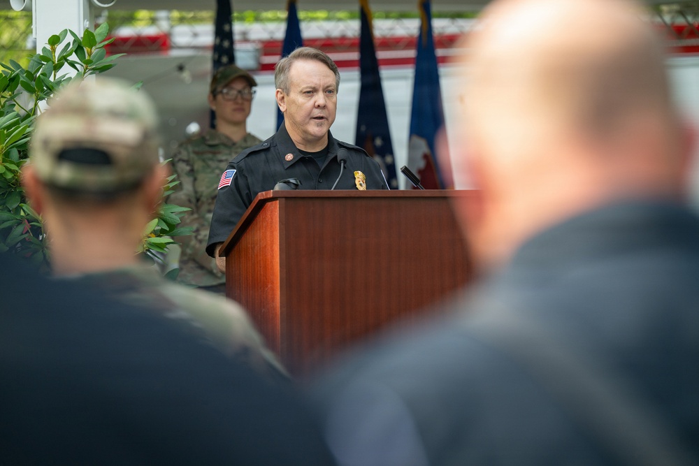 A uniformed official, appearing to be a fire department officer based on his black uniform with badge and American flag patch, is speaking at a wooden podium with a microphone. The event appears to be a formal ceremony or public address, with flags visible in the background and an audience in the foreground. A person in military camouflage uniform is visible standing behind the speaker.