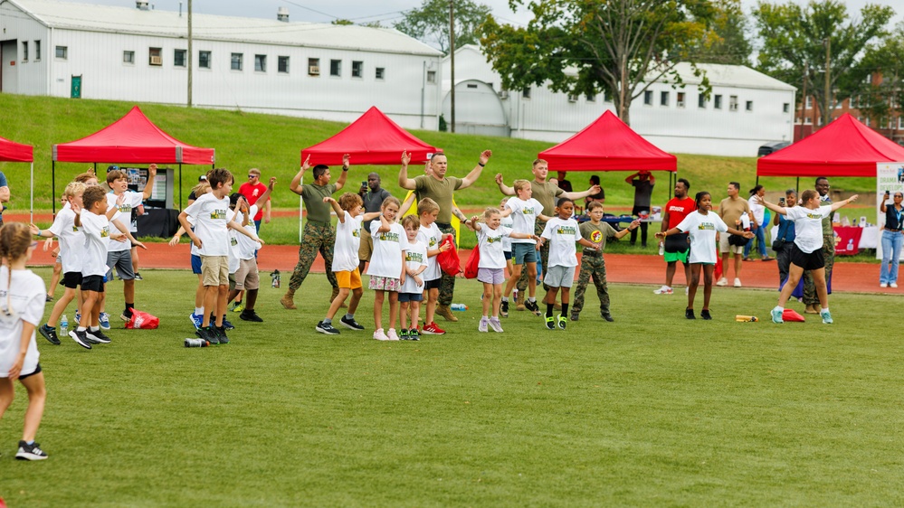 A group of children wearing matching white t-shirts participate in an outdoor sports or physical activity event on a green turf field, supervised by adults in military camouflage uniforms. Several red pop-up canopy tents are set up along the sidelines, suggesting an organized event or camp. The setting appears to be a military base or institutional facility based on the white buildings visible in the background.