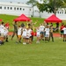 A group of children wearing matching white t-shirts participate in an outdoor sports or physical activity event on a green turf field, supervised by adults in military camouflage uniforms. Several red pop-up canopy tents are set up along the sidelines, suggesting an organized event or camp. The setting appears to be a military base or institutional facility based on the white buildings visible in the background.