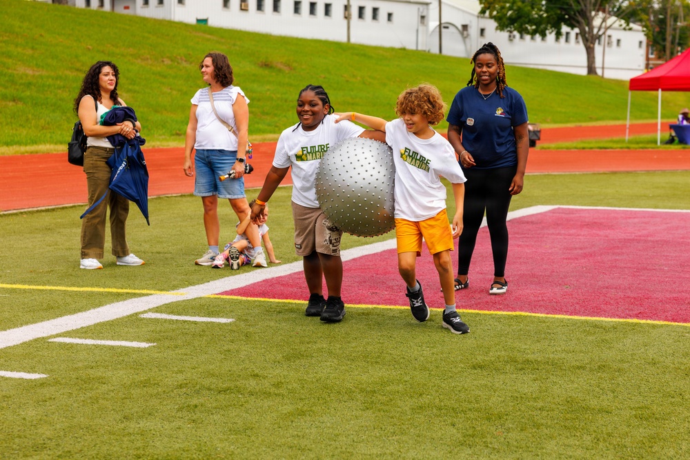 Two children wearing matching 'Future Marine' white t-shirts are participating in an outdoor activity on a turf athletic field, carrying a large silver exercise/stability ball together. They are supervised by an adult in a navy blue shirt, while other adults and a small child observe from the sideline. The event appears to be a youth program or camp activity held at a military or sports facility.