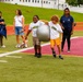 Two children wearing matching 'Future Marine' white t-shirts are participating in an outdoor activity on a turf athletic field, carrying a large silver exercise/stability ball together. They are supervised by an adult in a navy blue shirt, while other adults and a small child observe from the sideline. The event appears to be a youth program or camp activity held at a military or sports facility.