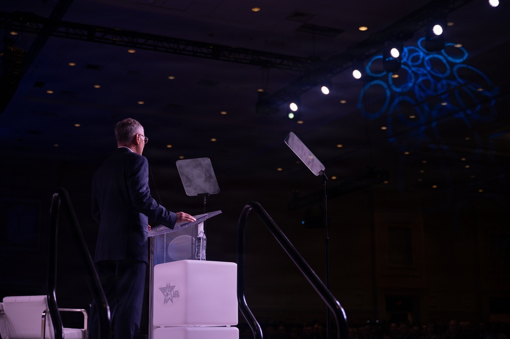 A man in a dark suit stands at a podium on a stage, appearing to deliver a speech or presentation at a formal conference or event. The podium features a logo that reads 'AFA' with a star emblem, suggesting an Air Force Association event. The large darkened venue features decorative blue lighting projections on the ceiling and stage lighting equipment.