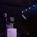 A man in a dark suit stands at a podium on a stage, appearing to deliver a speech or presentation at a formal conference or event. The podium features a logo that reads 'AFA' with a star emblem, suggesting an Air Force Association event. The large darkened venue features decorative blue lighting projections on the ceiling and stage lighting equipment.