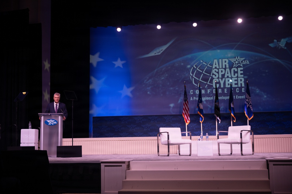 A man in a dark suit is speaking at a podium on a conference stage at the AFA Air Space Cyber Conference, described as 'A Professional Development Event.' The stage features multiple flags, two white chairs arranged for a panel discussion, and a large blue projection screen displaying the conference branding. The setting appears to be a large auditorium or convention hall.