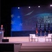 A man in a dark suit is speaking at a podium on a conference stage at the AFA Air Space Cyber Conference, described as 'A Professional Development Event.' The stage features multiple flags, two white chairs arranged for a panel discussion, and a large blue projection screen displaying the conference branding. The setting appears to be a large auditorium or convention hall.