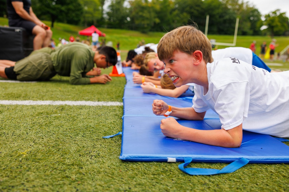 A group of children are lying prone on blue exercise mats on an artificial turf field during what appears to be an organized outdoor fitness or sports event. The foreground child, a boy in a white t-shirt with an orange wristband, displays intense determination with clenched fists and a grimacing expression. The scene suggests a competitive or timed physical challenge, likely a plank or low-crawl exercise.
