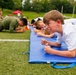 A group of children are lying prone on blue exercise mats on an artificial turf field during what appears to be an organized outdoor fitness or sports event. The foreground child, a boy in a white t-shirt with an orange wristband, displays intense determination with clenched fists and a grimacing expression. The scene suggests a competitive or timed physical challenge, likely a plank or low-crawl exercise.