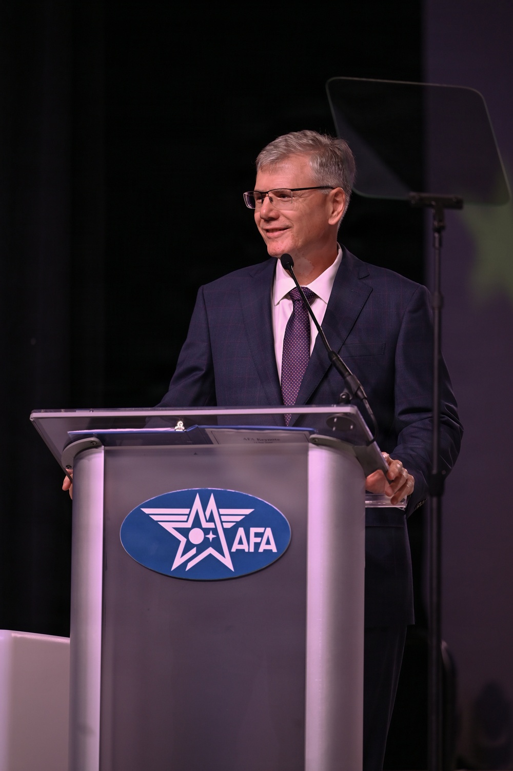 A middle-aged man with gray hair and glasses is speaking at a podium bearing the AFA (Air Force Association) logo. He is dressed in a navy blue suit with a purple patterned tie and appears to be delivering a speech or keynote address. The setting appears to be a formal conference or awards event.