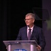 A middle-aged man with gray hair and glasses is speaking at a podium bearing the AFA (Air Force Association) logo. He is dressed in a navy blue suit with a purple patterned tie and appears to be delivering a speech or keynote address. The setting appears to be a formal conference or awards event.