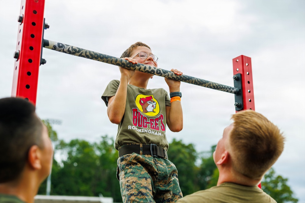 A young child wearing glasses, a Buc-ee's t-shirt, and military camouflage pants is performing pull-ups on a patterned bar mounted on a red pull-up rack. Two individuals, appearing to be military personnel, are observing and likely supervising the child. The setting appears to be an outdoor military recruitment or community engagement event.