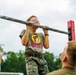 A young child wearing glasses, a Buc-ee's t-shirt, and military camouflage pants is performing pull-ups on a patterned bar mounted on a red pull-up rack. Two individuals, appearing to be military personnel, are observing and likely supervising the child. The setting appears to be an outdoor military recruitment or community engagement event.