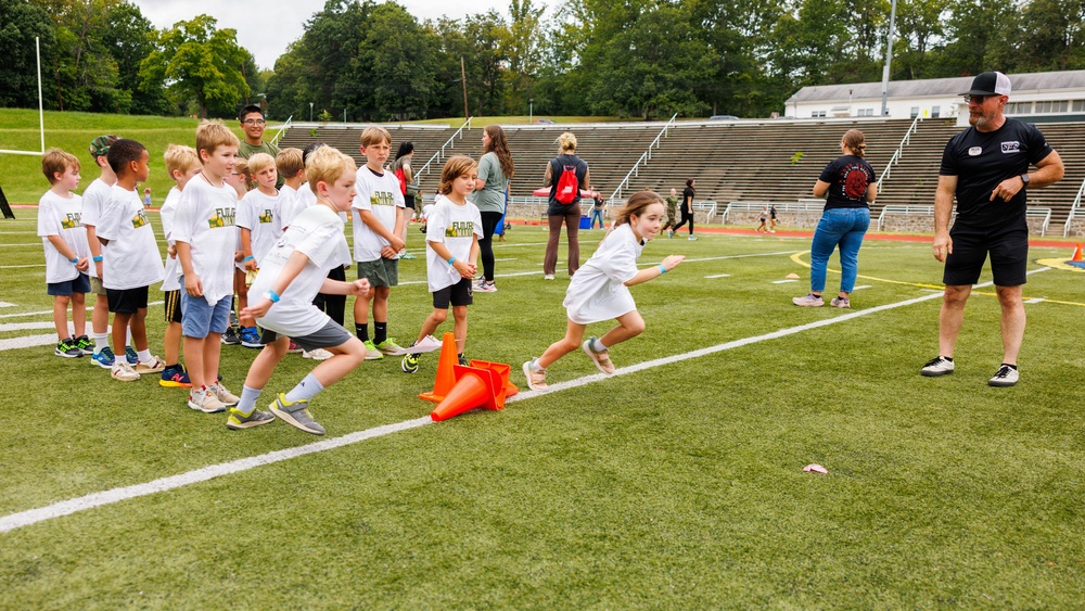 A youth sports camp or clinic is taking place on a football field inside a stadium. Children wearing matching white t-shirts with a logo are participating in a running or agility drill, starting from an orange cone marker. An adult coach in a black shirt and cap is observing and directing the activity.