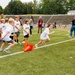 A youth sports camp or clinic is taking place on a football field inside a stadium. Children wearing matching white t-shirts with a logo are participating in a running or agility drill, starting from an orange cone marker. An adult coach in a black shirt and cap is observing and directing the activity.